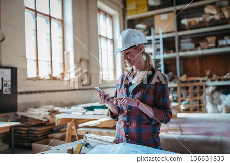 Female woodworker looking at email from client, standing in carpentry workshop. 136634833