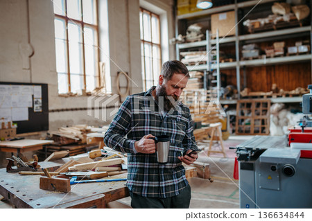 Woodworker looking at phone, standing in carpentry workshop. Woodworker looking at phone, standing in carpentry workshop. 136634844