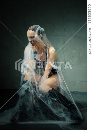Conceptual portrait of a woman ensnared in a cocoon of spiderwebs against a dark wall. Conceptual portrait of a woman ensnared in a cocoon of spiderwebs against a dark wall. 136635990