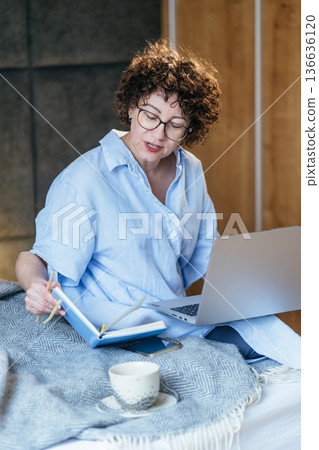 Business female engaged focused mood, laptop on bed, reaching for blue notebook with pencil, smartphone on blanket, scheduling week, productivity routine, home office 136636120
