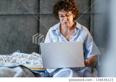 serious White woman in her 40s, curly hair, glasses, concentrated mood, using laptop on bed, home office routine, remote work, online tasks, journaling notebook nearby, productivity, digital lifestyle serious White woman in her 40s, curly hair, glasses, concentrated mood, using laptop on bed, home office routine, remote work, online tasks, journaling notebook nearby, productivity, digital lifestyle 136636123