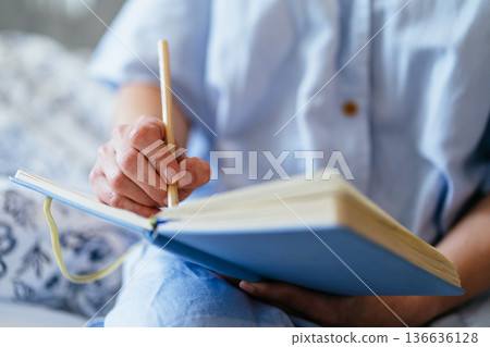 close up, middle aged White woman hand holding pencil, writing in blue notebook on bed, soft natural light, cozy home bedroom, mindful journaling, personal goals, quiet self care moment. close up, middle aged White woman hand holding pencil, writing in blue notebook on bed, soft natural light, cozy home bedroom, mindful journaling, personal goals, quiet self care moment. 136636128