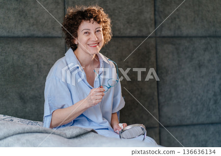 Charming woman with bouncy ringlets holding her glasses and smiling at the camera. Concept of successful morning grooming, self-confidence, and a happy, pensive start to a new day. Charming woman with bouncy ringlets holding her glasses and smiling at the camera. Concept of successful morning grooming, self-confidence, and a happy, pensive start to a new day. 136636134