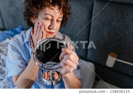 fashionable middle age 40s woman with defined curls focused on her facial skin detail. Concept of conscious grooming, aging with style, and a pensive, meticulous approach to beauty in a cozy interior. fashionable middle age 40s woman with defined curls focused on her facial skin detail. Concept of conscious grooming, aging with style, and a pensive, meticulous approach to beauty in a cozy interior. 136636135