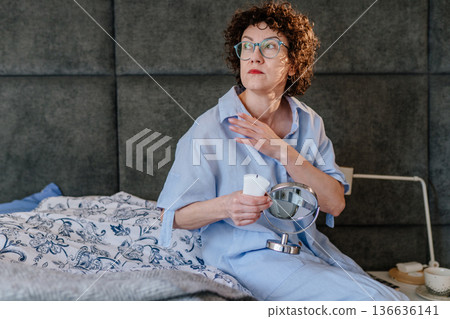 Reflective middle-aged woman in blue pajamas lost in thought while holding a cream tube. Concept of choosing skincare, aging gracefully, and pensive domestic moments in a cozy bedroom. Reflective middle-aged woman in blue pajamas lost in thought while holding a cream tube. Concept of choosing skincare, aging gracefully, and pensive domestic moments in a cozy bedroom. 136636141