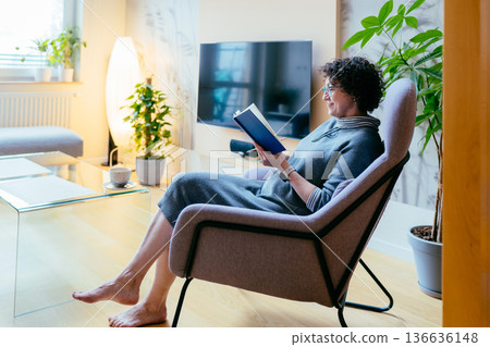 High angle of a mature woman in a blue armchair reading a book. Cozy home interior, hot tea, slow living and mindfulness concept. Mental health, education and peaceful weekend in modern apartment. High angle of a mature woman in a blue armchair reading a book. Cozy home interior, hot tea, slow living and mindfulness concept. Mental health, education and peaceful weekend in modern apartment. 136636148
