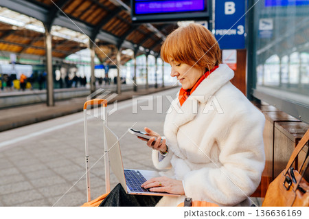 Experienced caucasian redhead professional using smartphone and laptop at train station platform. Managing communication and tasks during travel, hybrid work routine, in public transport environment. Experienced caucasian redhead professional using smartphone and laptop at train station platform. Managing communication and tasks during travel, hybrid work routine, in public transport environment. 136636169