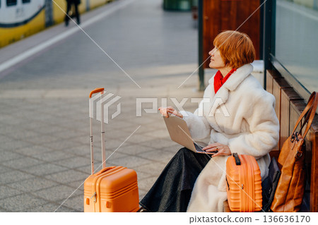 Thoughtful middle aged redhead woman sitting on railway station platform with laptop and suitcase. Before decision, professional experience, independence and control during winter urban transit. Thoughtful middle aged redhead woman sitting on railway station platform with laptop and suitcase. Before decision, professional experience, independence and control during winter urban transit. 136636170