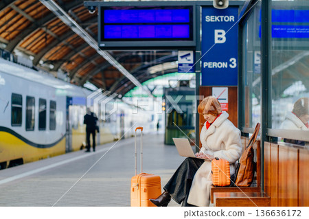 Confident middle aged redhead woman using laptop on railway station platform with travel suitcase. Experienced organizing transit, responsibility independent career in winter city transport. Confident middle aged redhead woman using laptop on railway station platform with travel suitcase. Experienced organizing transit, responsibility independent career in winter city transport. 136636172