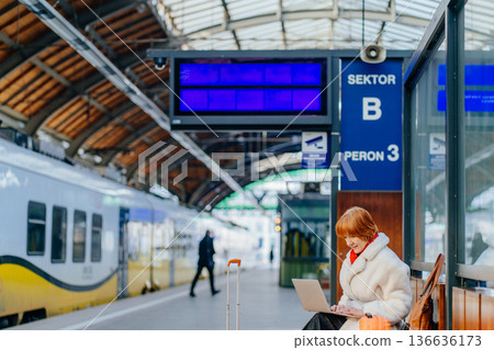 Successful middle aged redhead woman sitting on bench at railway station platform with suitcase and laptop. Work during travel, combining business routine, mobility in winter urban transport . 136636173