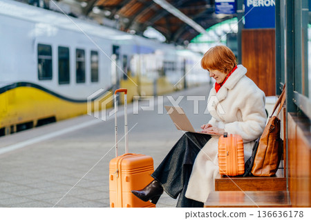 Caucasian redhead woman working on laptop while waiting for train at railway station platform. Freelance lifestyle, remote work, mobility, travel, modern work routine during winter journey. 136636178