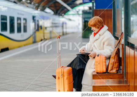 Caucasian redhead adult woman sitting on wooden bench at railway station, using smartphone, waiting for train, orange suitcase, white coat, red scarf, winter indoor platform , solo travel routine 136636180