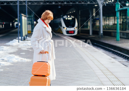 Caucasian redhead adult woman standing on railway platform with orange suitcase, white coat and red scarf, looking down with calm focused expression, electric train, winter urban rail travel moment Caucasian redhead adult woman standing on railway platform with orange suitcase, white coat and red scarf, looking down with calm focused expression, electric train, winter urban rail travel moment 136636184