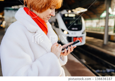 Close view of caucasian redhead adult woman holding smartphone on railway station platform, white coat and red scarf, checking information before train arrival, digital travel routine Close view of caucasian redhead adult woman holding smartphone on railway station platform, white coat and red scarf, checking information before train arrival, digital travel routine 136636187