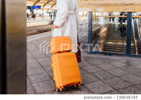 Rear view of woman walking on railway station platform, pulling orange suitcase on wheels, white coat, travel by train, everyday rail commute, eco friendly public transport, urban space Rear view of woman walking on railway station platform, pulling orange suitcase on wheels, white coat, travel by train, everyday rail commute, eco friendly public transport, urban space 136636193