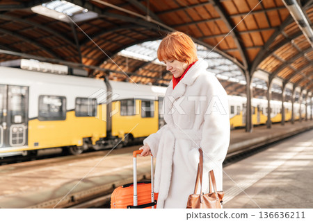 Caucasian adult woman on railway station platform near yellow train, holding orange suitcase, leather handbag, wearing white fur coat red scarf, looking down, winter travel, eco friendly transport 136636211