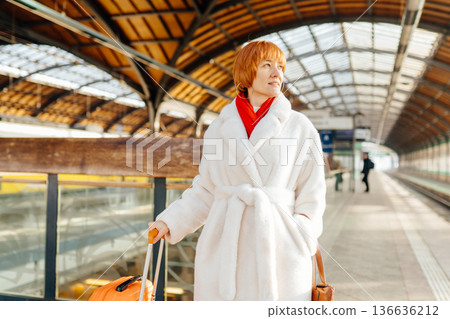 Caucasian redhead adult woman standing on railway station platform, with orange suitcase, white fur coat, red scarf, looking aside, winter travel, mobility, eco transport, journey, departure moment Caucasian redhead adult woman standing on railway station platform, with orange suitcase, white fur coat, red scarf, looking aside, winter travel, mobility, eco transport, journey, departure moment 136636212