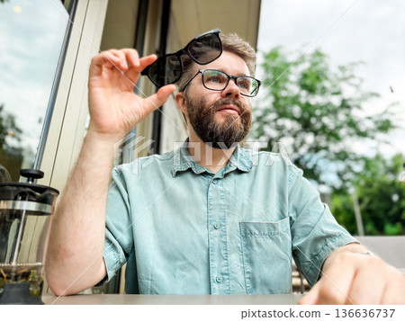 Glasses with magnetic clip on sunglasses. Man smiling while wearing clip on sunglasses at outdoor cafe table Glasses with magnetic clip on sunglasses. Man smiling while wearing clip on sunglasses at outdoor cafe table 136636737