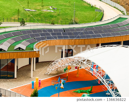 Building with solar panels installed on curved roof near green landscape and playground. Renewable energy, technology, and sustainability symbolizing innovation, ecology, and environmental balance. Building with solar panels installed on curved roof near green landscape and playground. Renewable energy, technology, and sustainability symbolizing innovation, ecology, and environmental balance. 136636822