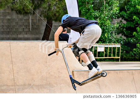 Young man practicing Scootering (Freestyle Scootering) in the new SkatePark in the central park of Igualada, Barcelona, Spain. Young man practicing Scootering (Freestyle Scootering) in the new SkatePark in the central park of Igualada, Barcelona, Spain. 136636905