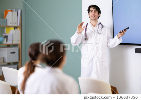 Portrait of young man doctor standing near interactive board and showing presentation with remote control for group of medicals students sitting in chairs in auditorium 136637350