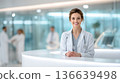 Female doctor in white coat smiles at reception desk in modern medical facility with blurred colleagues in background, copy space available 136639498