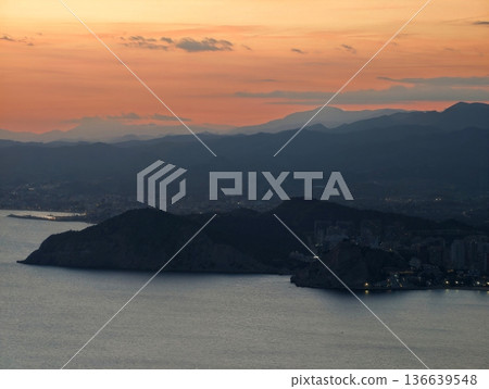 Dramatic Silhouette Of Headland Against Sunset Sky With Peaceful Bay And Tiny City Lights Along Shoreline Dramatic Silhouette Of Headland Against Sunset Sky With Peaceful Bay And Tiny City Lights Along Shoreline 136639548