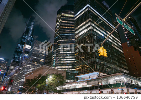 Tall glass skyscrapers tower above East 53rd Street in New York City, illuminated by office lights and traffic signals. A yellow streetlight and dark sky add contrast to the vibrant urban night scene. Tall glass skyscrapers tower above East 53rd Street in New York City, illuminated by office lights and traffic signals. A yellow streetlight and dark sky add contrast to the vibrant urban night scene. 136640759