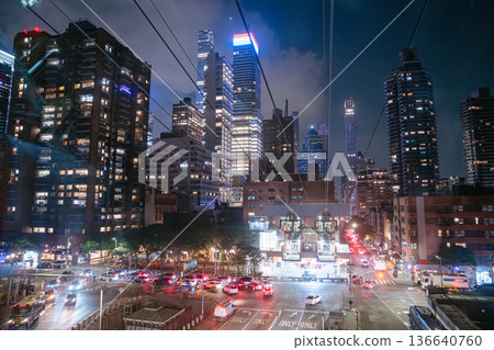 The Roosevelt Island Tram prepares for departure above a busy Midtown street filled with traffic and lights. Skyscrapers and glowing windows create a vibrant nighttime cityscape in New York City. The Roosevelt Island Tram prepares for departure above a busy Midtown street filled with traffic and lights. Skyscrapers and glowing windows create a vibrant nighttime cityscape in New York City. 136640760