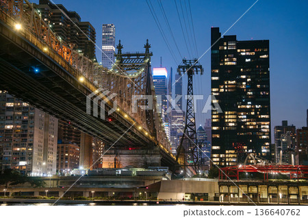 A detailed view of the Queensboro Bridge shows glowing lights and steel structure beside the Roosevelt Island Tram tower. Tall skyscrapers of Midtown Manhattan rise in the background during twilight. 136640762