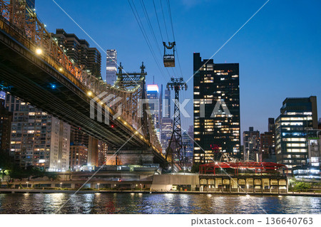 The illuminated Queensboro Bridge stretches over the East River with the Roosevelt Island Tram gliding above the city. Skyscrapers light up the background, creating a vibrant and dynamic New York The illuminated Queensboro Bridge stretches over the East River with the Roosevelt Island Tram gliding above the city. Skyscrapers light up the background, creating a vibrant and dynamic New York 136640763