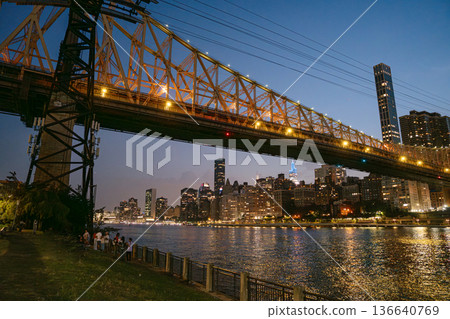 The Queensboro Bridge stretches over the East River as the Manhattan skyline glows in the background. A group of people stands by the waterfront, enjoying the city lights and evening atmosphere. The Queensboro Bridge stretches over the East River as the Manhattan skyline glows in the background. A group of people stands by the waterfront, enjoying the city lights and evening atmosphere. 136640769