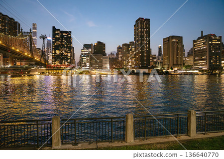 The New York City skyline shines with illuminated skyscrapers beside the Queensboro Bridge at twilight. The river reflects golden lights, and a quiet metal fence lines the calm waterfront in the The New York City skyline shines with illuminated skyscrapers beside the Queensboro Bridge at twilight. The river reflects golden lights, and a quiet metal fence lines the calm waterfront in the 136640770