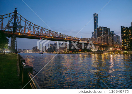 The Queensboro Bridge glows with lights as it stretches over the East River, connecting to Manhattan's vibrant skyline. The illuminated buildings and bridge reflect beautifully on the calm evening 136640775
