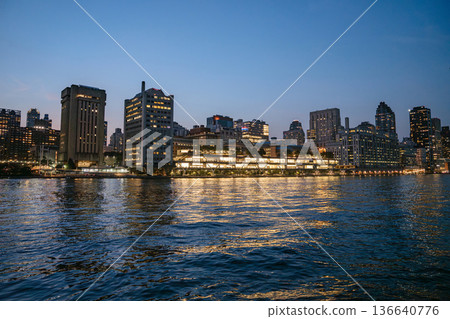 Modern office and residential buildings along the Manhattan waterfront glow brightly against the night sky. Their lights shimmer across the surface of the East River, creating a vibrant city 136640776