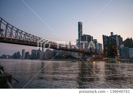 The Queensboro Bridge spans across the East River as Manhattan's illuminated skyline reflects in the calm water. The scene captures the peaceful beauty of the city during the last light of day. The Queensboro Bridge spans across the East River as Manhattan's illuminated skyline reflects in the calm water. The scene captures the peaceful beauty of the city during the last light of day. 136640781