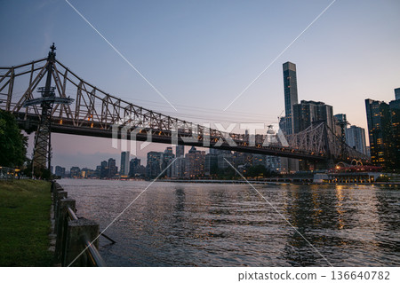 The Queensboro Bridge stretches across the East River as the Manhattan skyline lights up in the evening. Viewed from the riverside path, the scene captures the calm water and towering buildings under 136640782