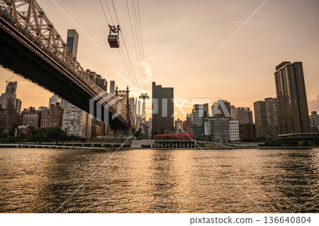 The Roosevelt Island tram glides above the East River with the Queensboro Bridge beside it. A warm sunset casts golden light over the Manhattan skyline and shimmering water. 136640804