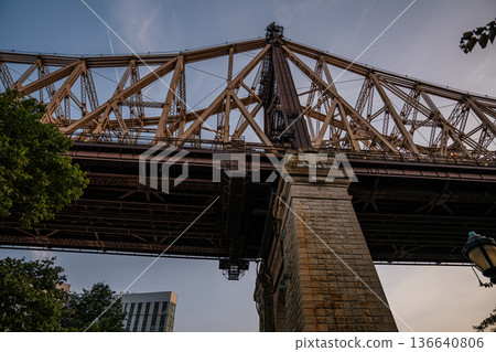 A dramatic upward view of the Queensboro Bridge captured during the evening light. The intricate steel framework and stone pillar highlight the bridge's architectural detail. A dramatic upward view of the Queensboro Bridge captured during the evening light. The intricate steel framework and stone pillar highlight the bridge's architectural detail. 136640806