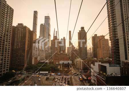 A dramatic view of the Manhattan skyline framed by tramway wires at sunset. Skyscrapers glow in warm light as traffic moves below in the heart of the city. A dramatic view of the Manhattan skyline framed by tramway wires at sunset. Skyscrapers glow in warm light as traffic moves below in the heart of the city. 136640807