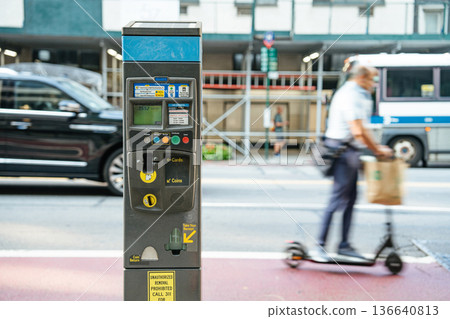 A parking meter stands on a New York City street as a man rides a scooter past it. The blurred motion of the rider contrasts with the stationary meter and busy urban background. A parking meter stands on a New York City street as a man rides a scooter past it. The blurred motion of the rider contrasts with the stationary meter and busy urban background. 136640813