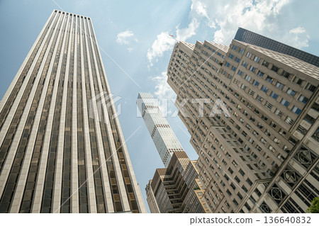 Towering skyscrapers rise above the streets of Midtown Manhattan under a bright summer sky. The modern glass buildings contrast with historic architecture in this iconic New York City view. Towering skyscrapers rise above the streets of Midtown Manhattan under a bright summer sky. The modern glass buildings contrast with historic architecture in this iconic New York City view. 136640832