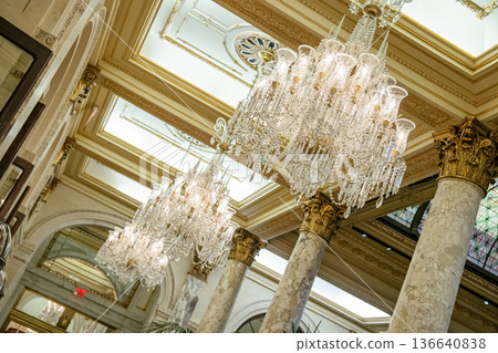Sparkling crystal chandeliers hang from an ornate gold-trimmed ceiling above marble columns in an upscale historic building. The elegant architecture reflects timeless grandeur and refined detail. 136640838