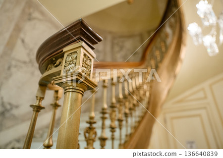 A close-up of an ornate golden stair railing features a floral carving and polished wooden handrail. The elegant curved staircase in the background glows under a crystal chandelier. 136640839