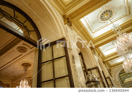 An elegant hall showcases arched wooden doors, intricate gold ceiling details, and glowing crystal chandeliers. The classic architectural elements and marble columns create a luxurious, timeless 136640844