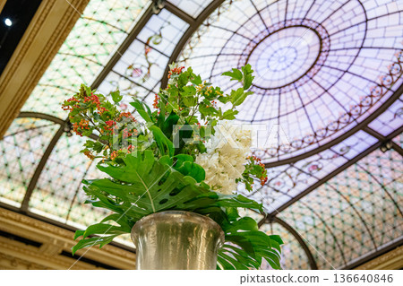 A luxurious flower arrangement stands in a tall silver vase beneath a stained glass dome ceiling. Bright green leaves, white blossoms, and red berries add vivid color to the ornate indoor setting. A luxurious flower arrangement stands in a tall silver vase beneath a stained glass dome ceiling. Bright green leaves, white blossoms, and red berries add vivid color to the ornate indoor setting. 136640846