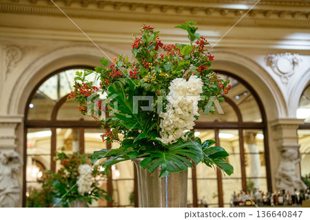 A grand silver vase holds a lush floral arrangement of white hydrangeas, red berries, and large tropical leaves. The vibrant display decorates the classic interior of a historic New York hotel with 136640847