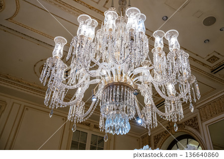 A detailed view of an ornate crystal chandelier shines brightly from the ceiling of a historic luxury hotel. Glass droplets, intricate shapes, and elegant lighting showcase opulence and craftsmanship 136640860