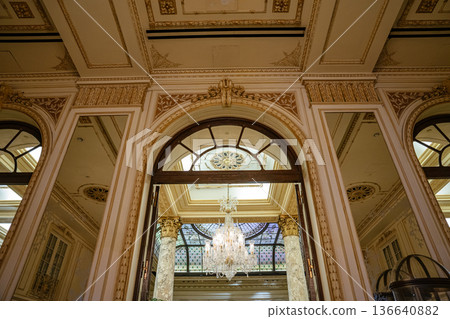 An ornate arched doorway opens to a grand chandelier and stained glass ceiling framed by marble columns. Detailed moldings, gold accents, and architectural symmetry reflect classic luxury in a An ornate arched doorway opens to a grand chandelier and stained glass ceiling framed by marble columns. Detailed moldings, gold accents, and architectural symmetry reflect classic luxury in a 136640882
