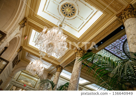 Elegant crystal chandeliers hang from a richly detailed ceiling with golden trim and classical molding. The luxurious hotel interior features marble columns, arched doorways, and stained glass panels. 136640949